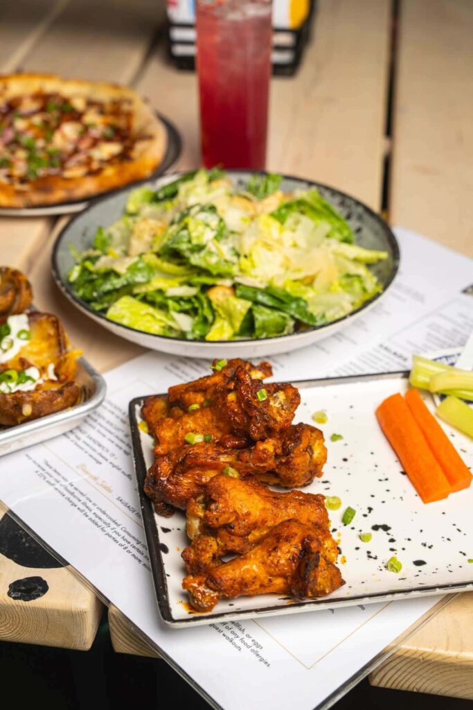 A close-up of buffalo chicken wings served with celery, carrots, and dipping sauce, accompanied by a Caesar salad, pizza, and a red cocktail.