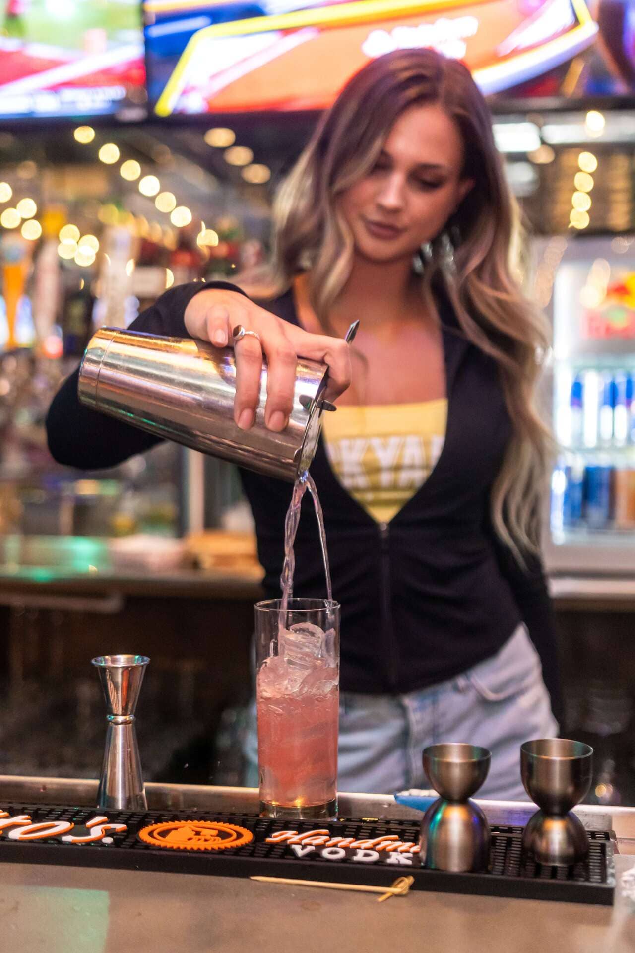 Bartender pours a pink cocktail over ice at a modern bar, with TV screens and cocktail tools in the background.