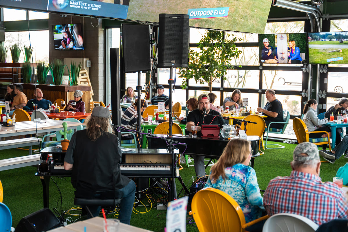 Indoor restaurant scene with live dueling piano performance and diners enjoying food at colorful tables.