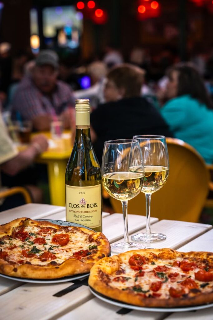 Two personal pizzas and a bottle of Clos du Bois Chardonnay served with wine glasses on a table at a busy restaurant.