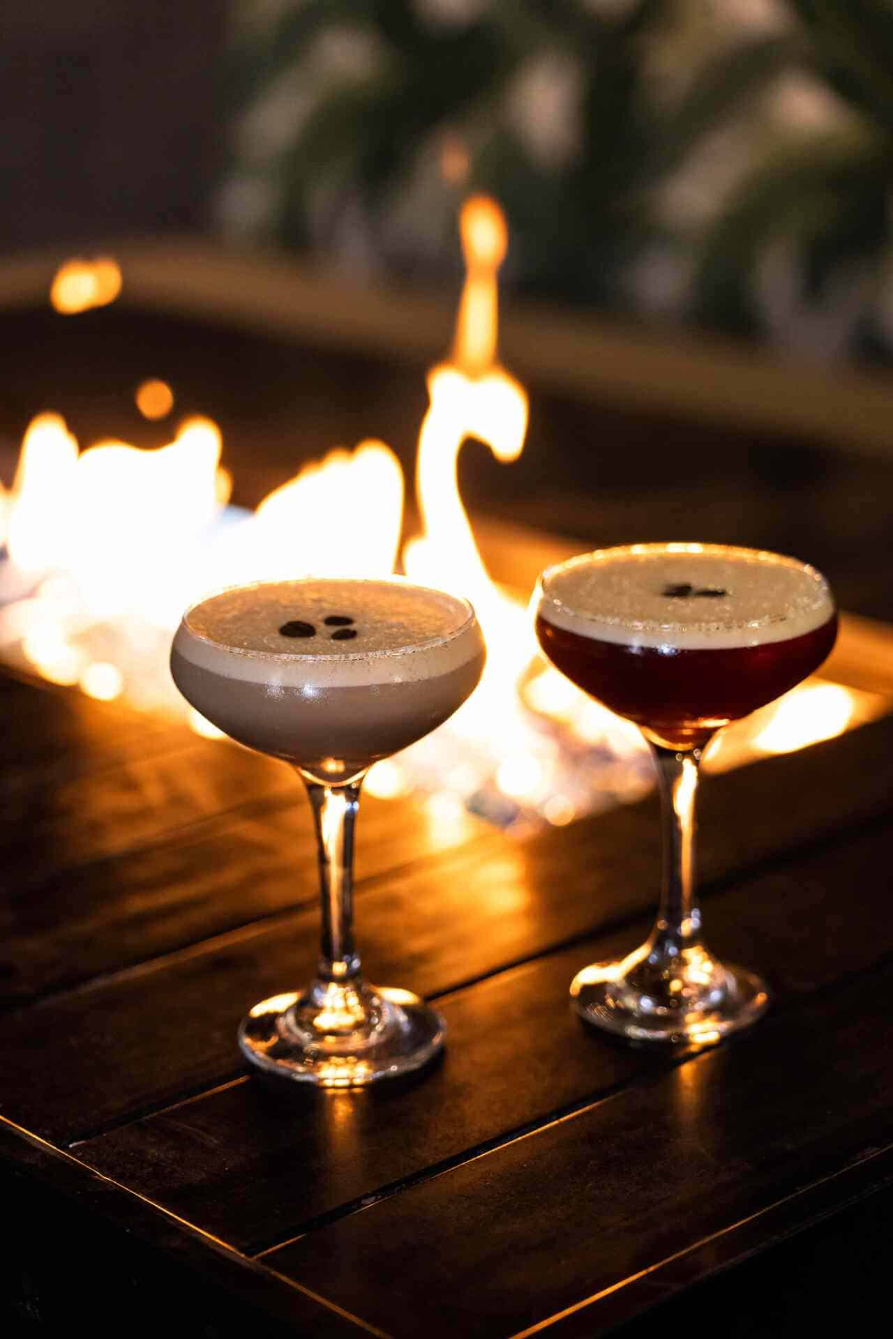 Two espresso martinis on a dark wooden table in front of a fire pit, with coffee beans as garnish and a blurred warm background.