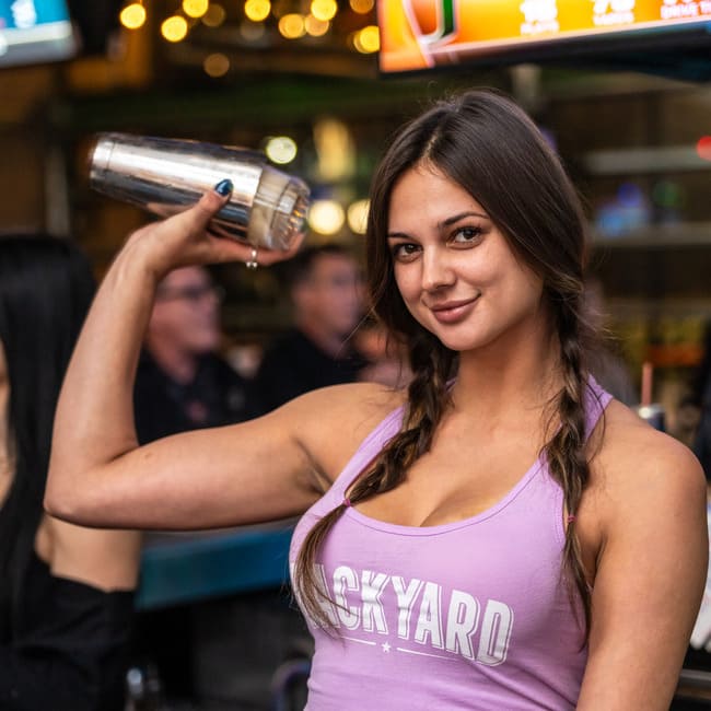 Woman in a lavender tank top smiling while holding a metal cocktail shaker behind her head at a bar.