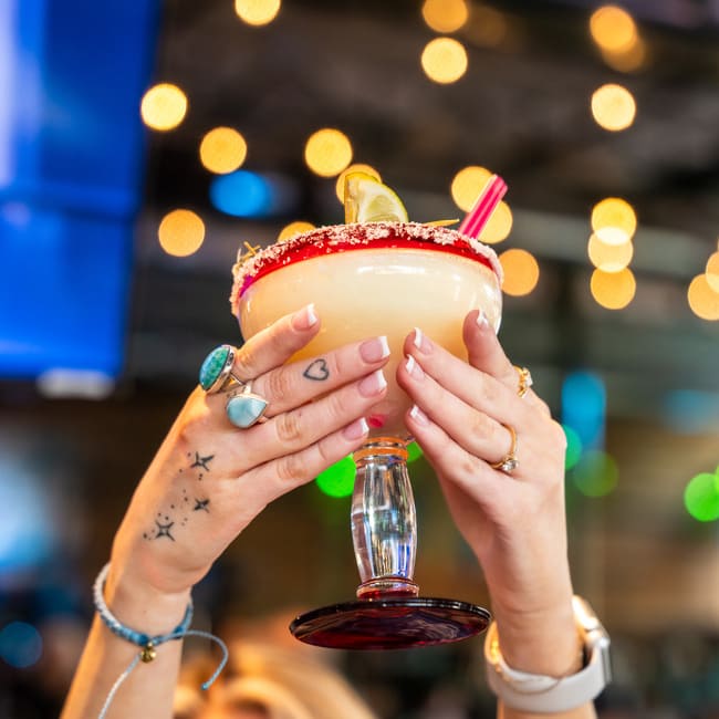 Hands holding a margarita cocktail with a salted rim and lime garnish against blurred bar lights.