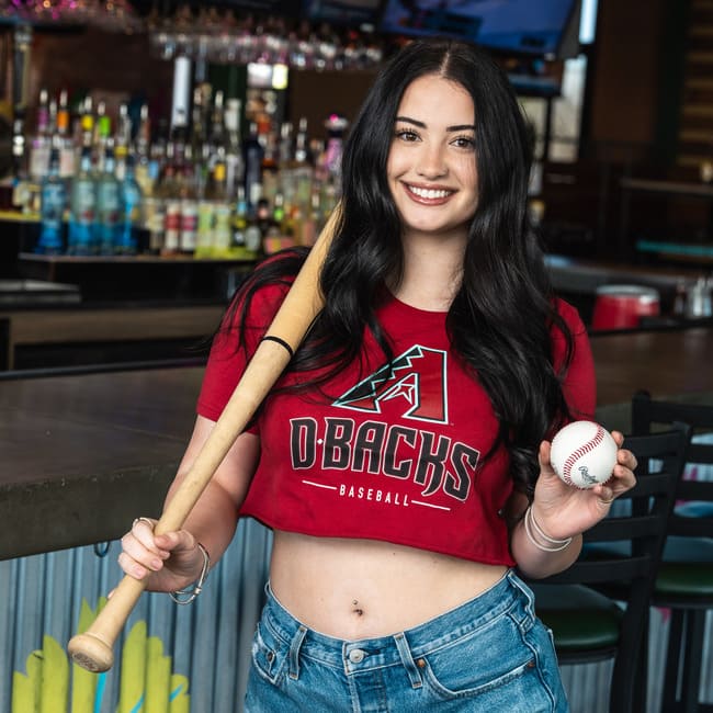 Woman holding a baseball bat and ball wearing a Dbacks shirt inside a bar