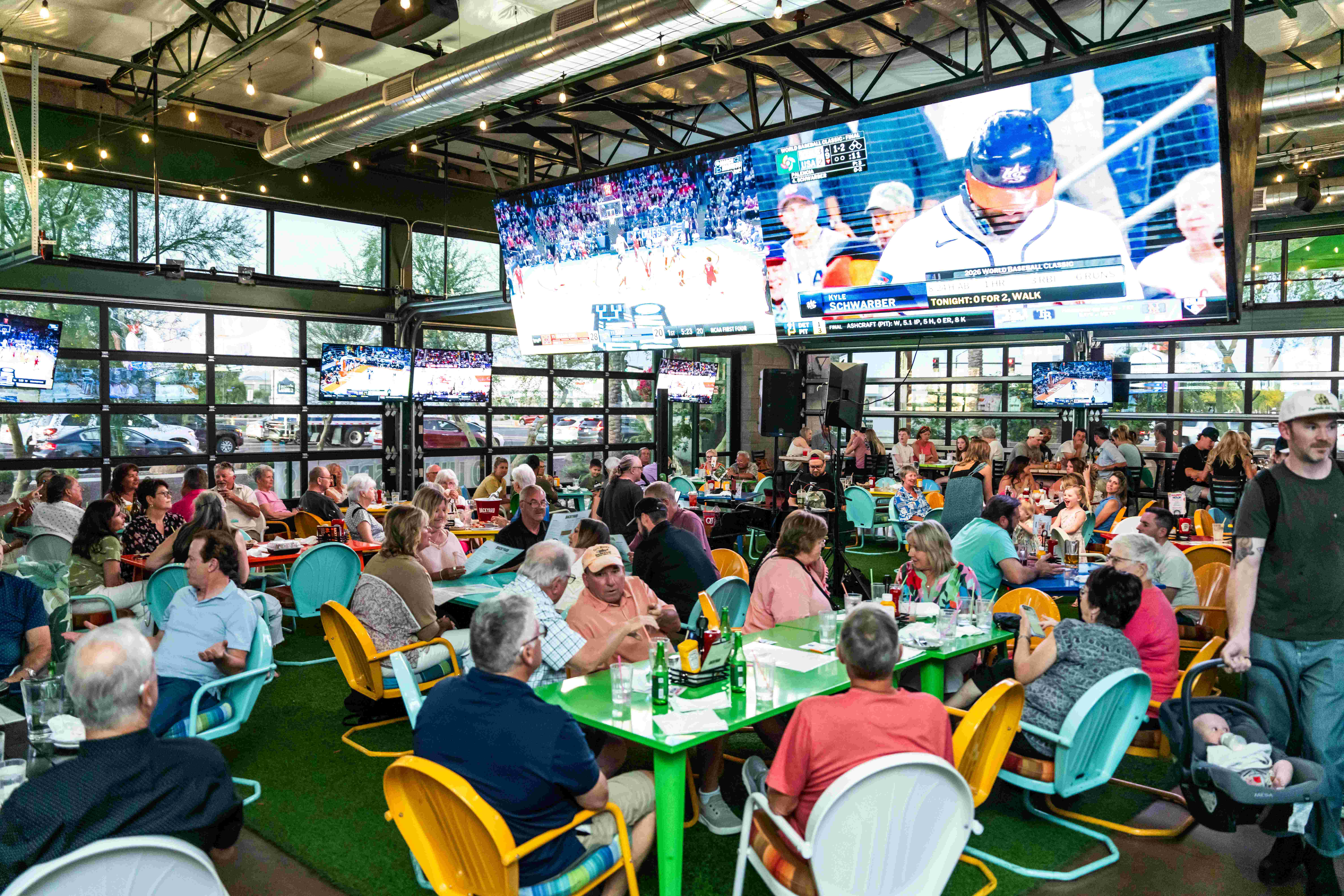 Busy indoor restaurant with large TV screens showing sports games and a crowd of people dining at colorful tables.