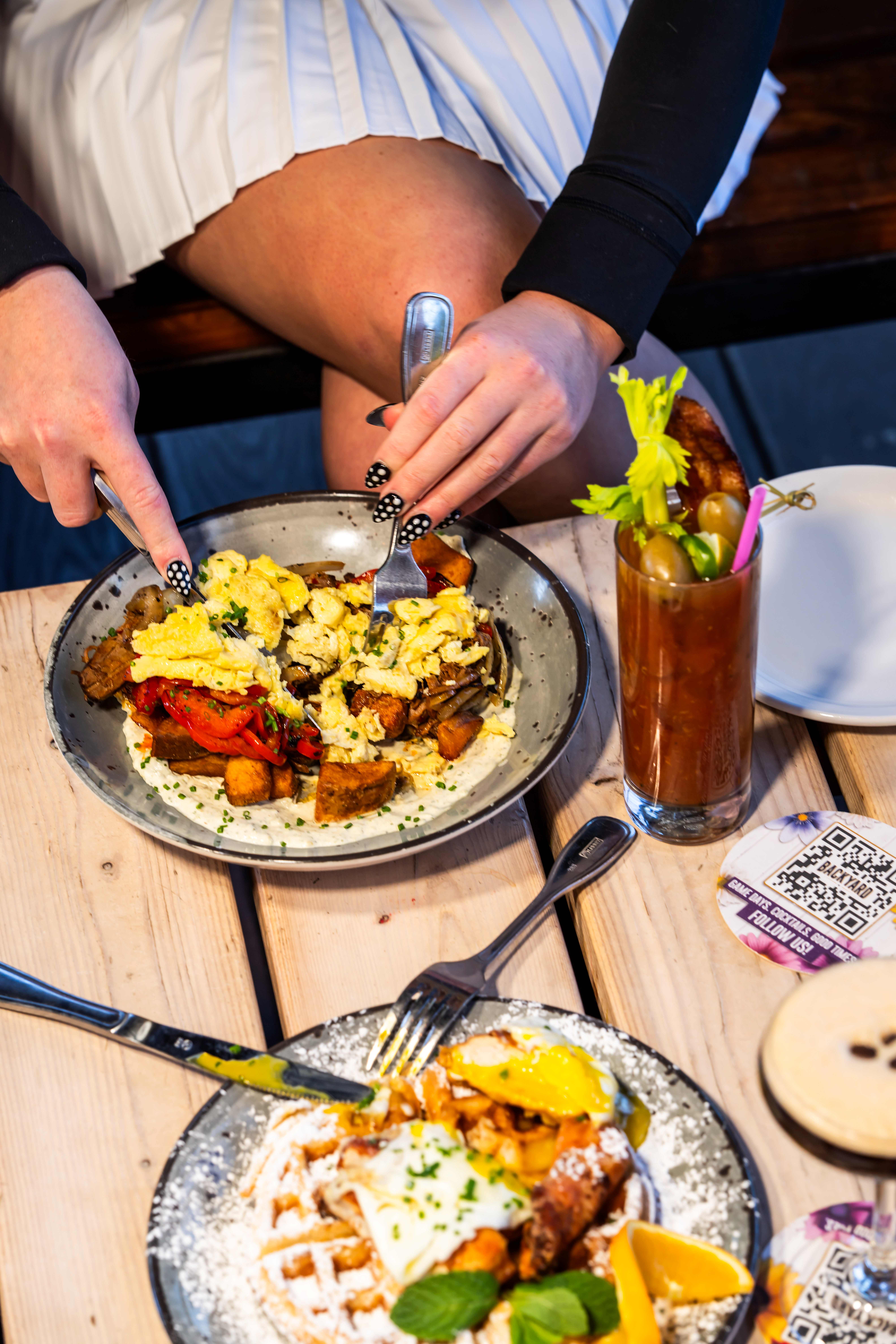 Brunch dishes with scrambled eggs, potatoes, and waffles topped with eggs, served alongside a Bloody Mary cocktail on a wooden table.