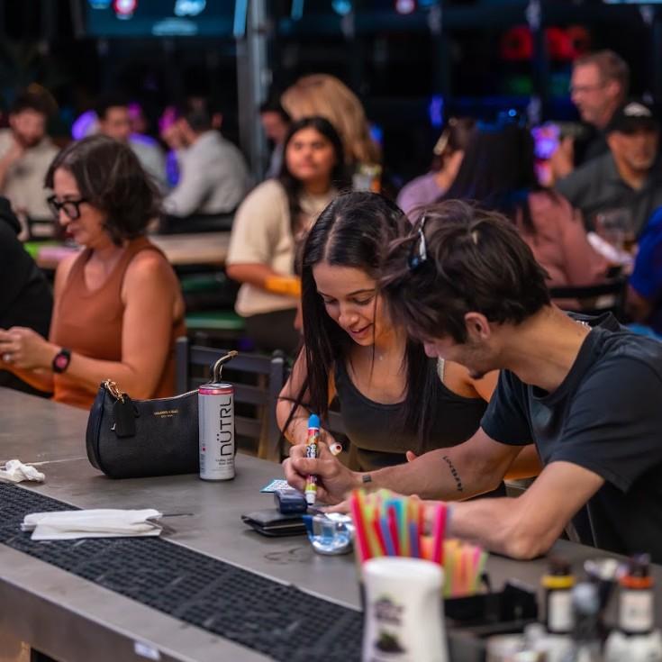 Two friends fill out bingo cards while enjoying a comedy bingo event at a lively sports bar, with other guests socializing in the background.