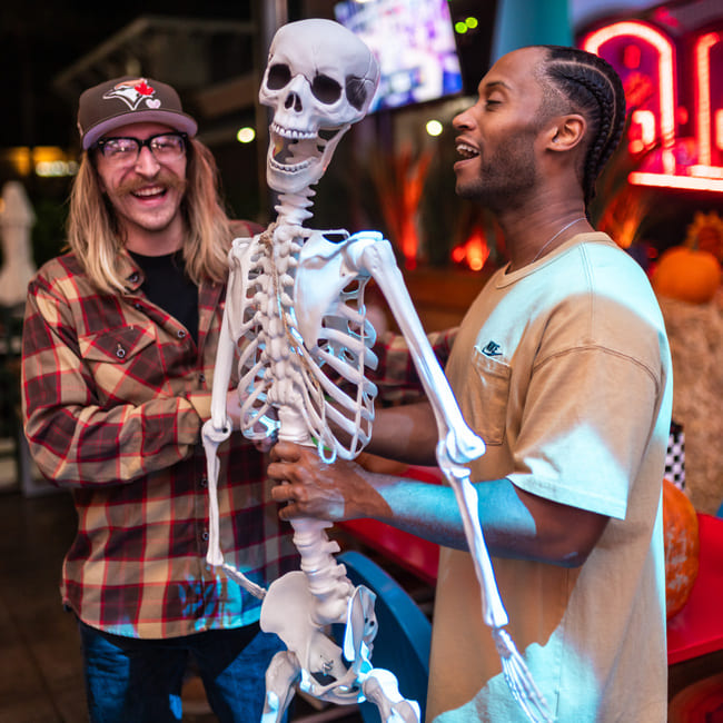 Two guests laughing while posing with a decorative skeleton during a Halloween celebration at Backyard.
